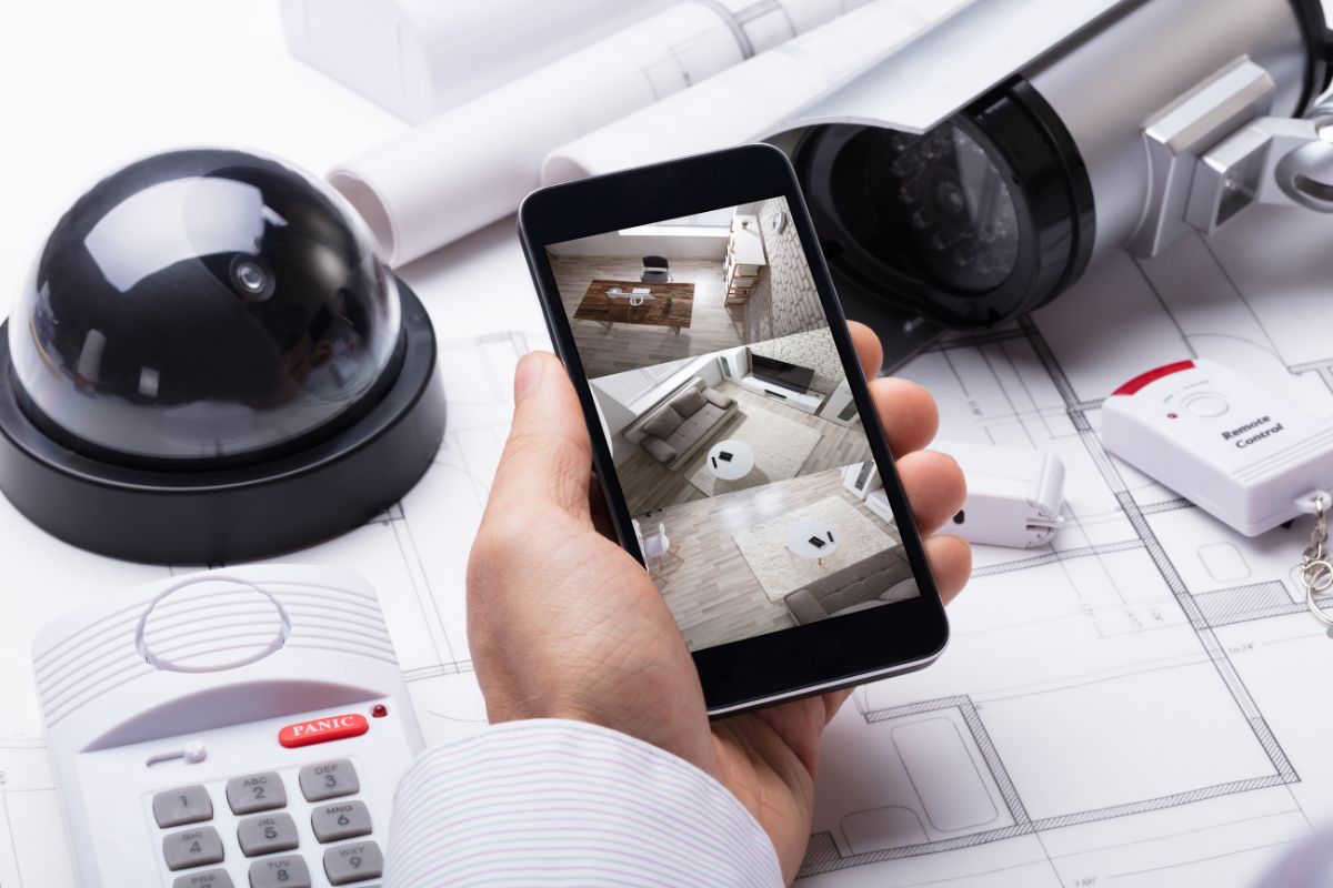  A person holds a smartphone displaying a camera app next to a mounted security camera on a wall.